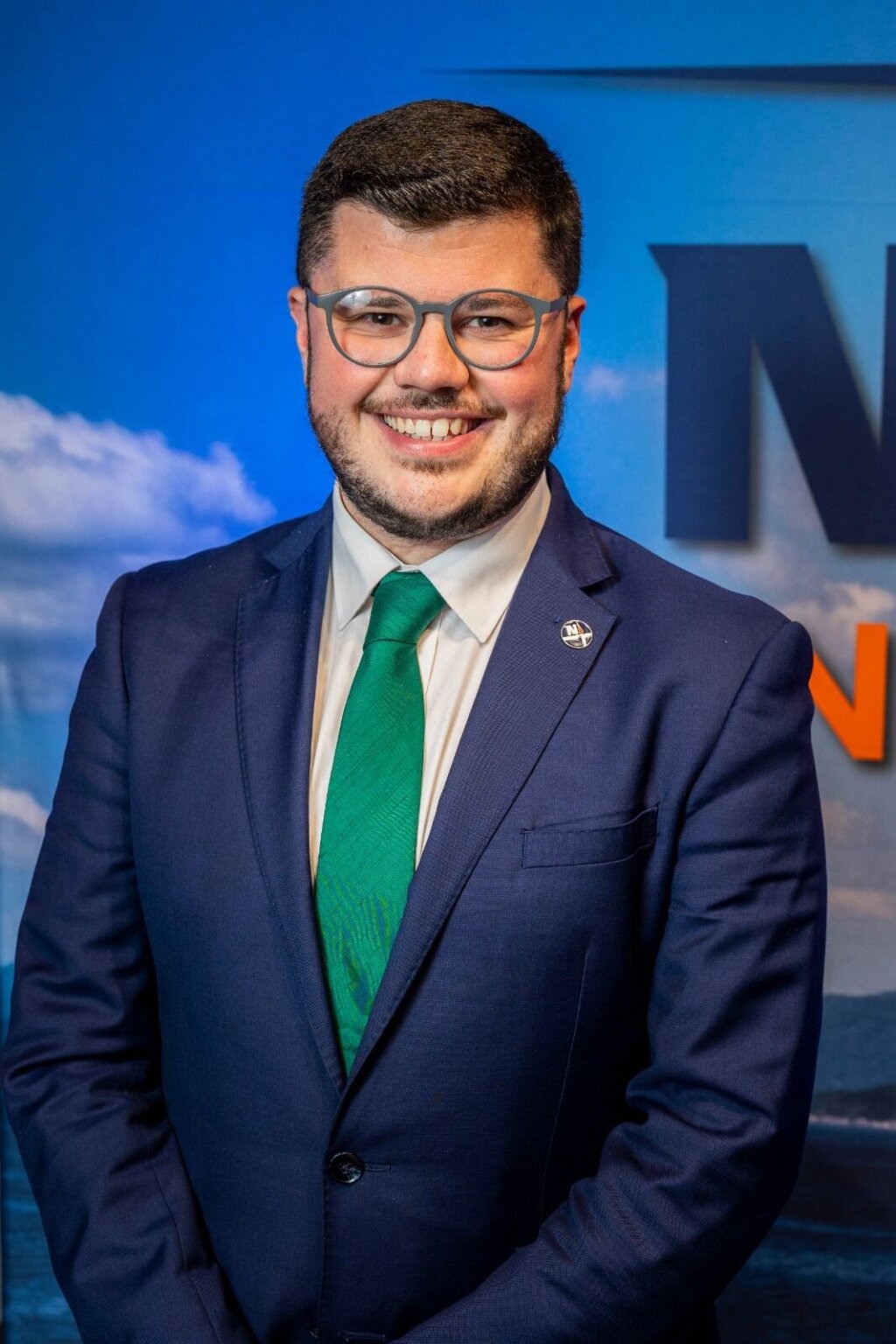A picture of a smiling man, here is wearing a navy suit and green tie.  He has dark hair and wears glasses.